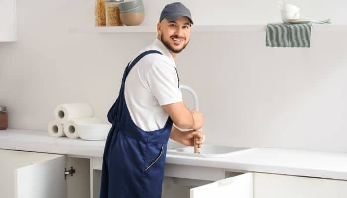 Plumber wearing blue overalls clearing a drain in a kitchen.